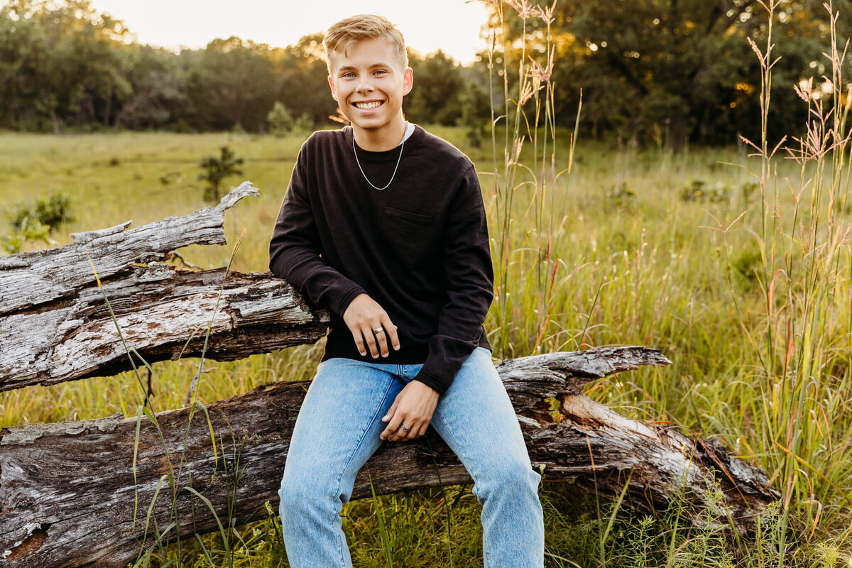 handsome teen boy sitting on a log in a field at sunset by Ashley Kalbus