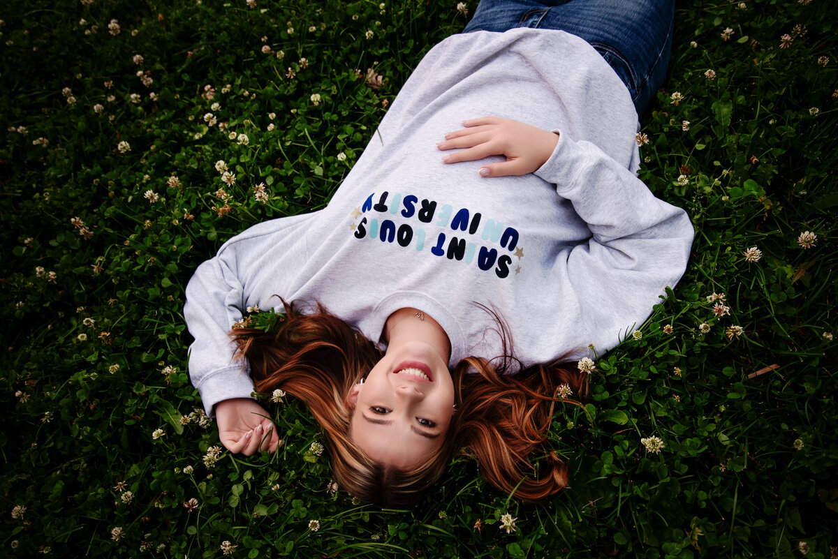 st-louis-senior-photographer-senior-girl-laying-in-grass-wearing-st-louis-university-sweatshirt