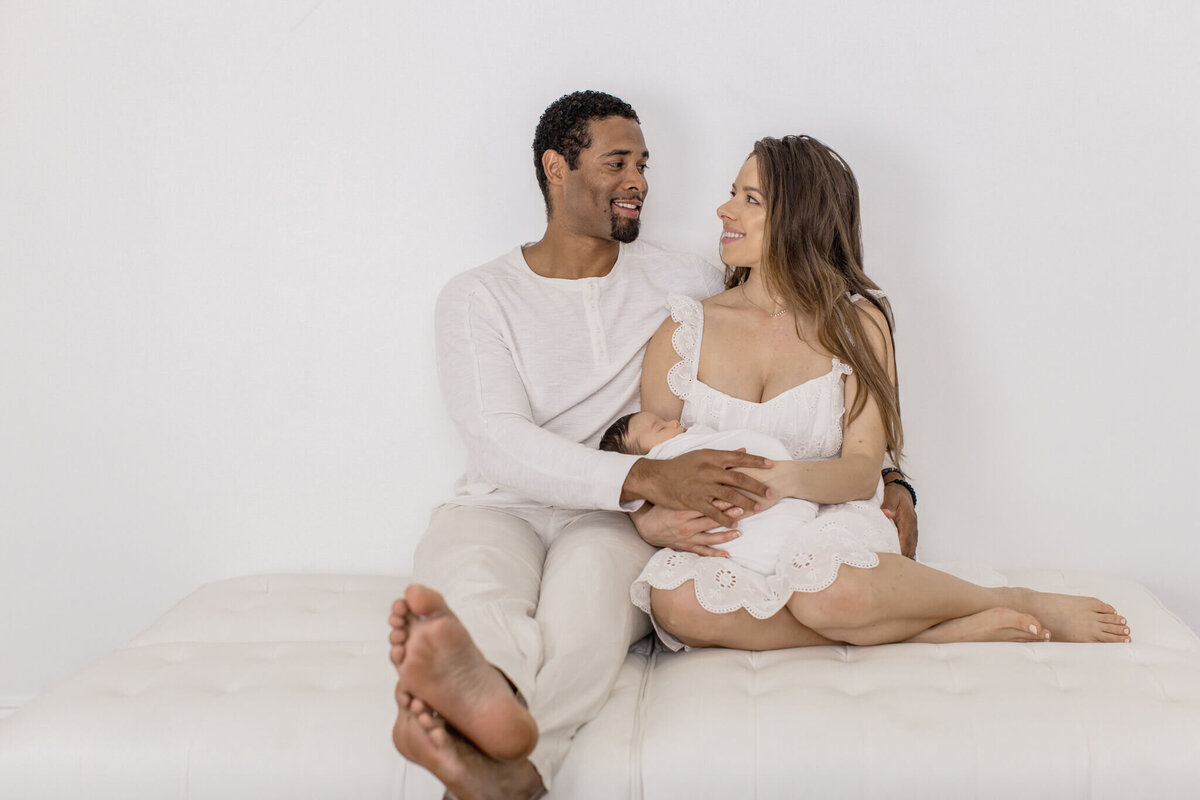 A couple sits closely on a white bed, both dressed in white. They are smiling and looking at each other, conveying a sense of affection and warmth. The background is a plain white wall, enhancing the serene and intimate atmosphere.