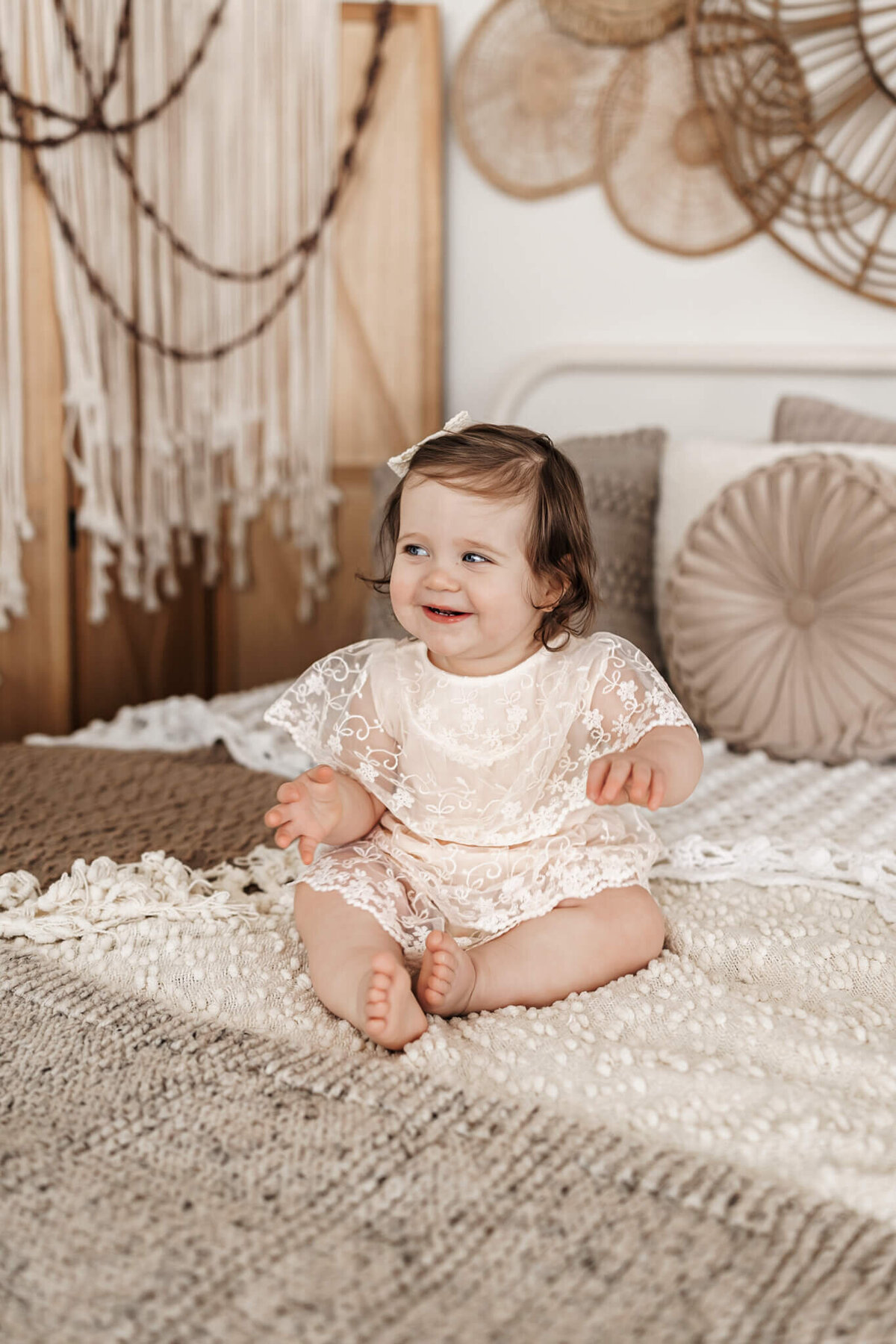 A smiling baby in a lacy, cream-colored outfit sits on a cozy, textured bed with neutral pillows. Woven decor and a green plant decorate the stylish, boho-inspired bedroom—perfect for milestone celebration portraits.