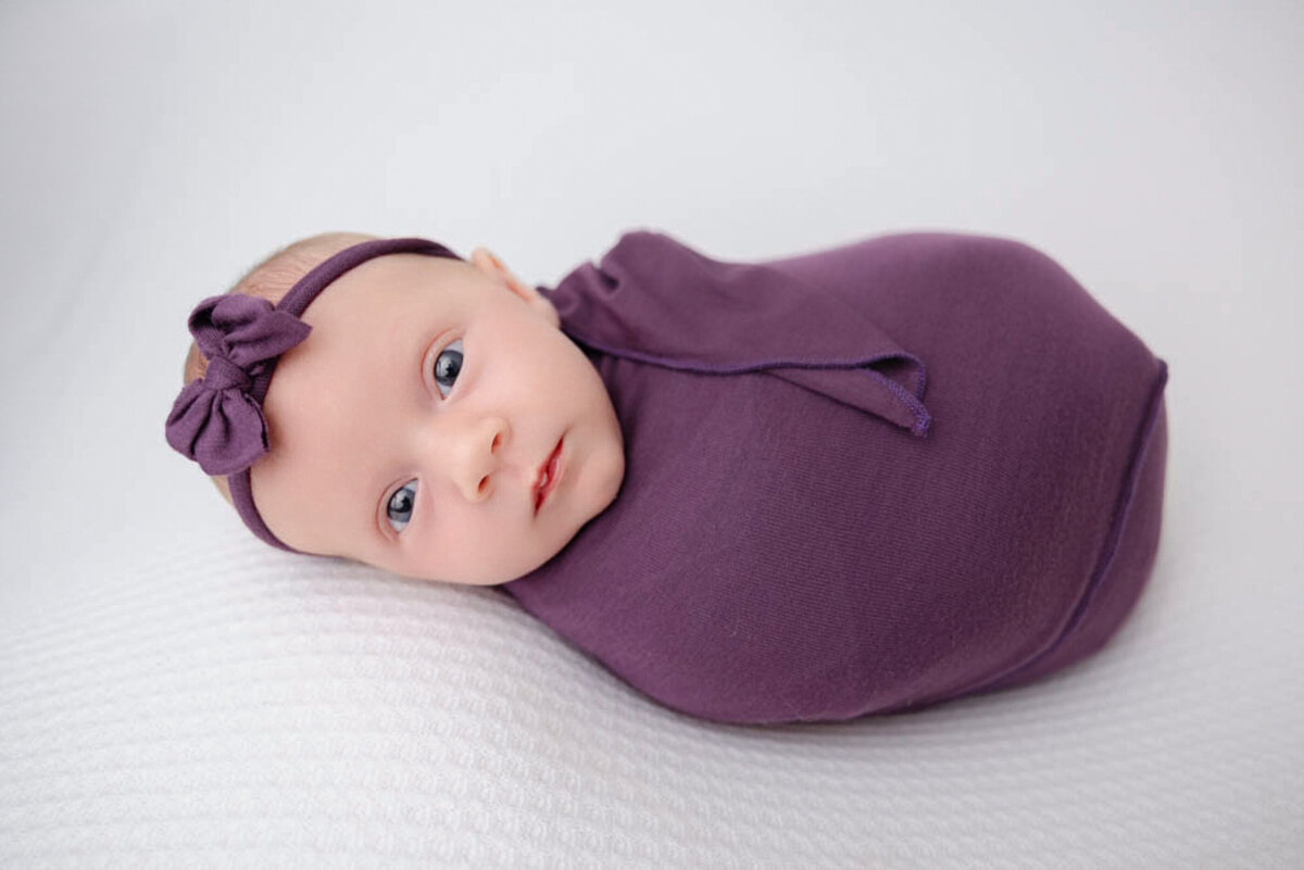 two-week baby girl posed lying on white blanket and wearing a deep purple head bow and wrap.