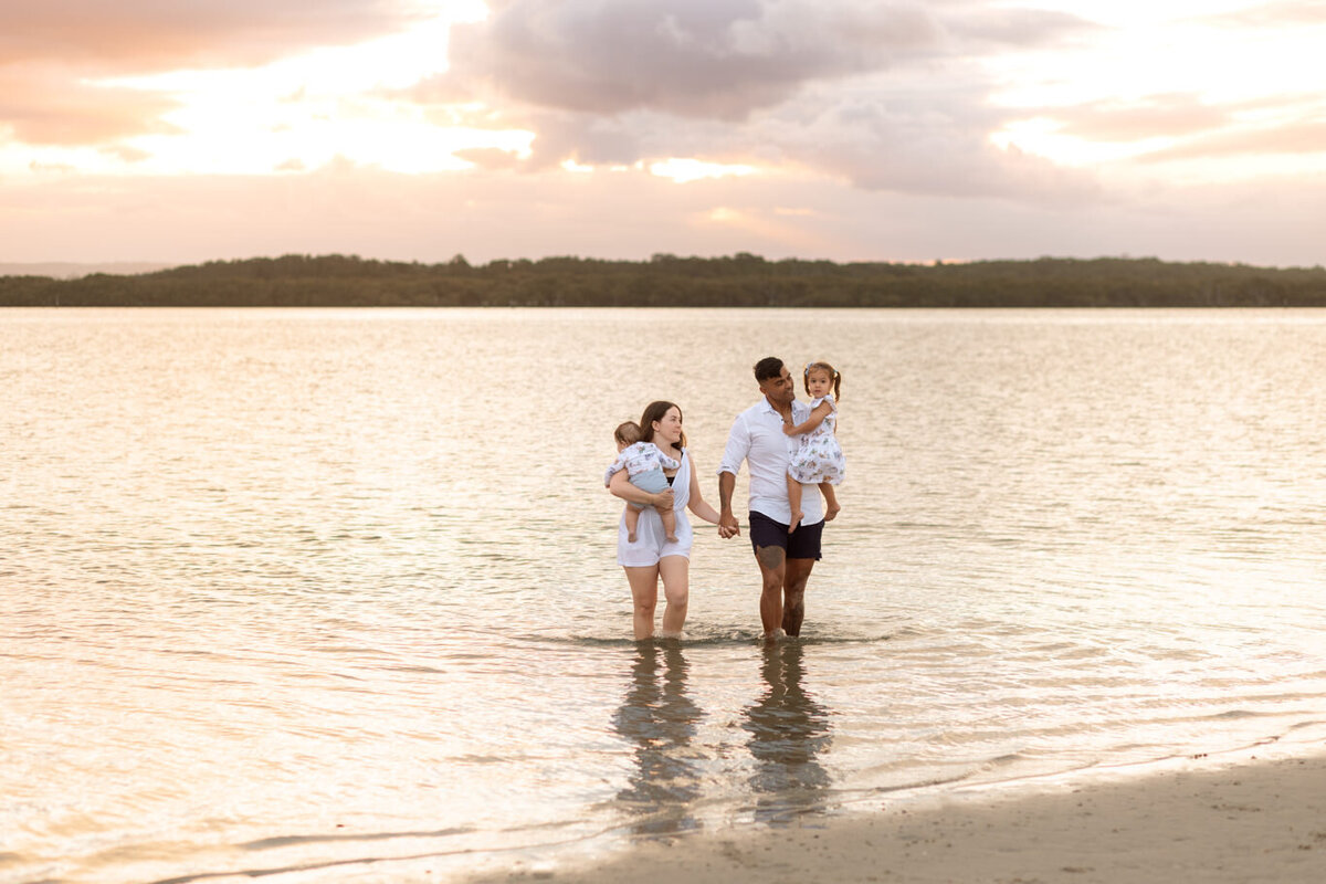 Family of four walking through shallow water at sunset during a warm and relaxed beach photo session in Sydney, captured in soft golden light.