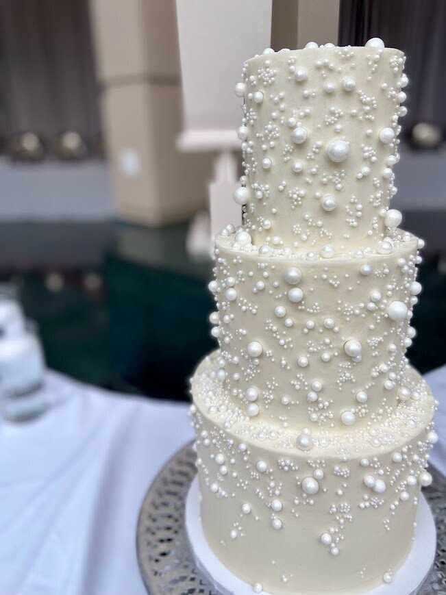 Close up shot of a three tier cake with white frosting. Scattered all over the cake are different sizes of sugar pearls, with more concentrated toward the top tier, and tapering off at the bottom tier.