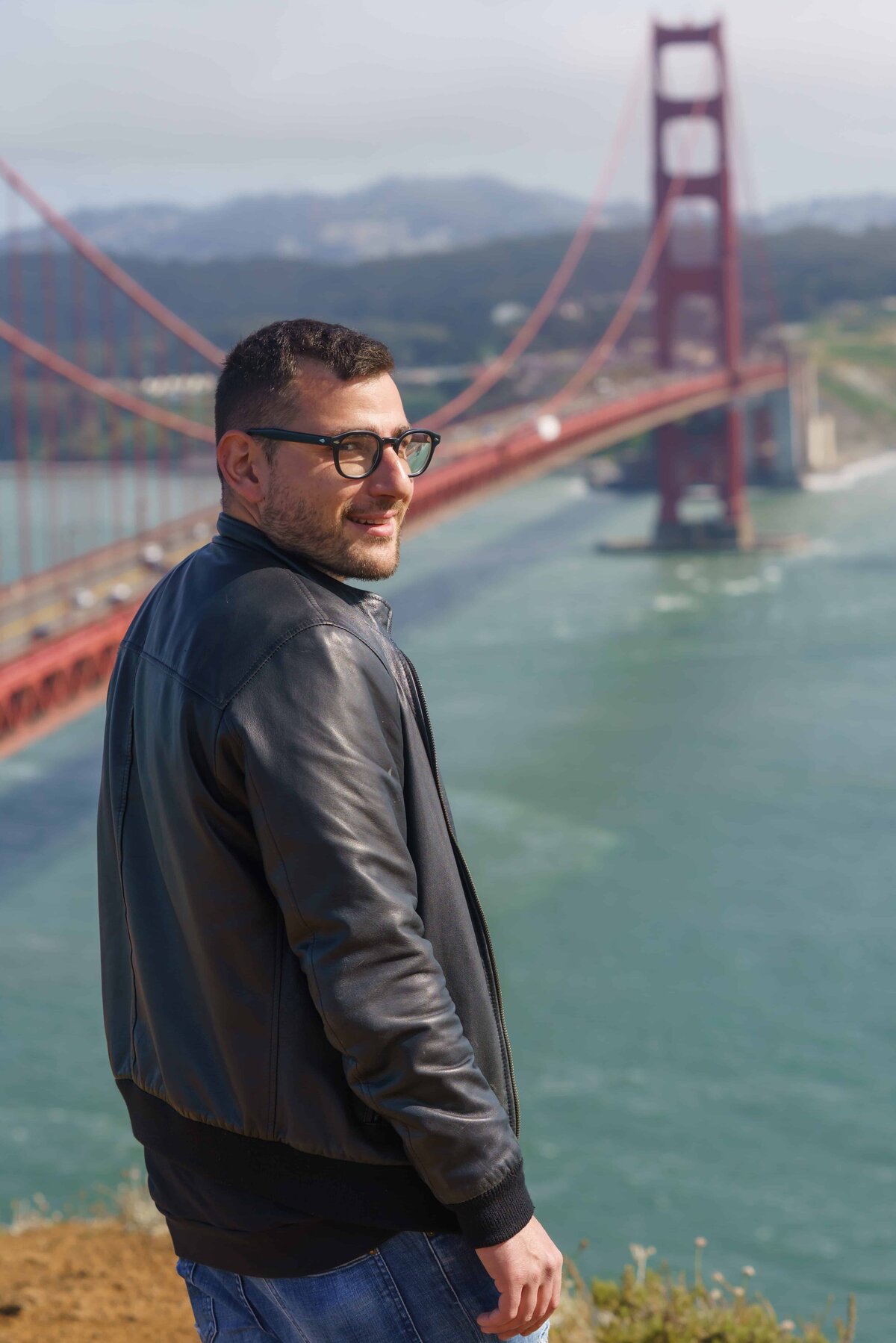 Man with a black leather jacket and glasses standing on a cliff overlooking the golden gate bridge