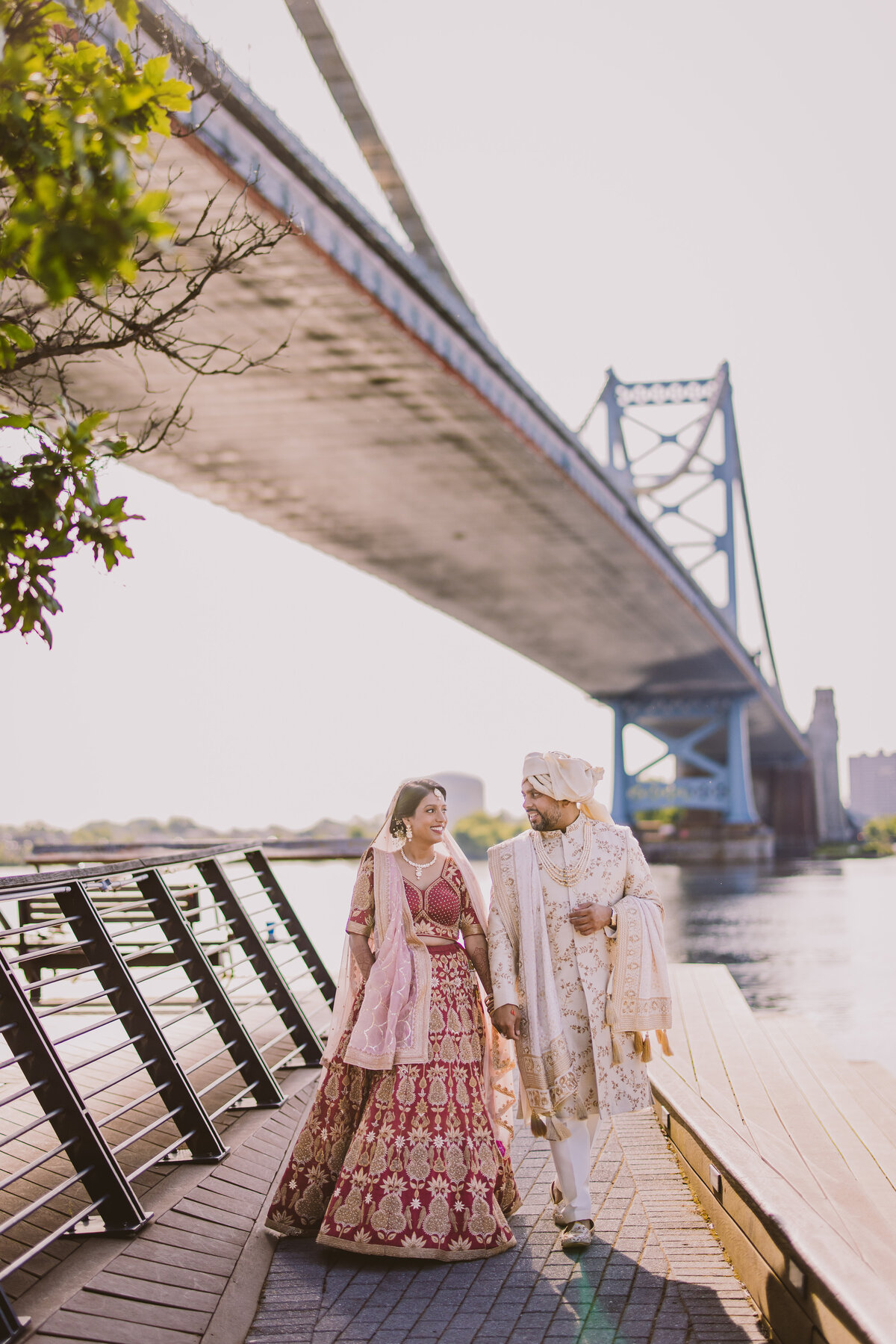 Indian bride and groom walking down the race street pier with the benjamin franklin bridge behind them