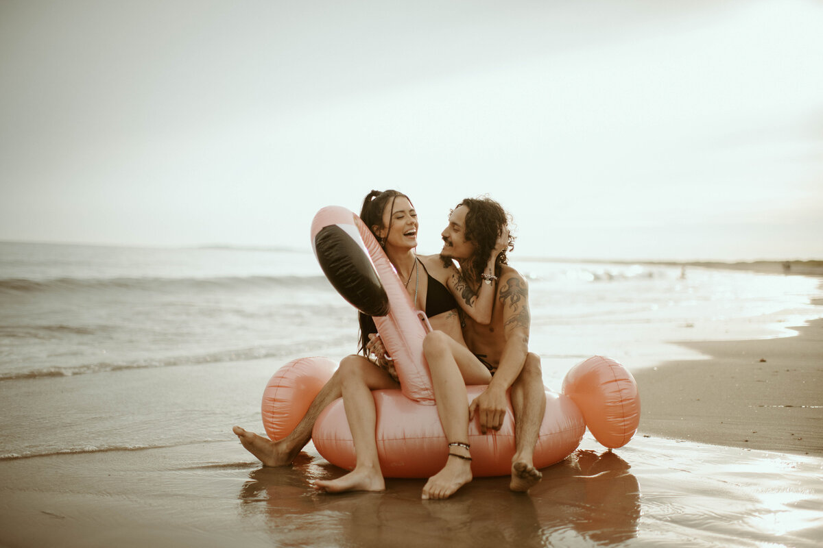 playful couple sits on beached flamingo raft in watch hill, RI