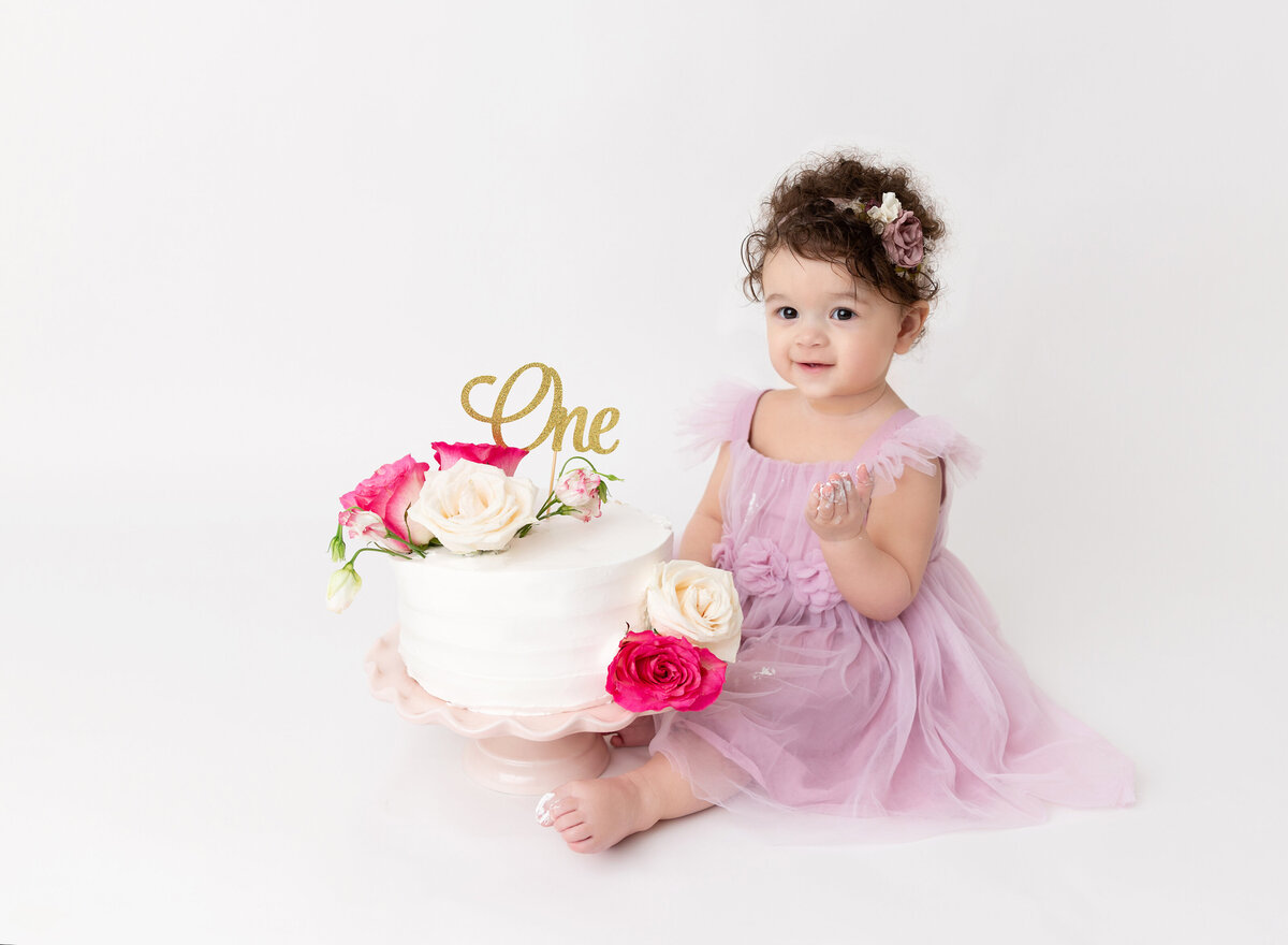 Baby girl in a lilac organza dress sits in front of a white cake decorated with fresh flowers. Baby is smiling at the camera.