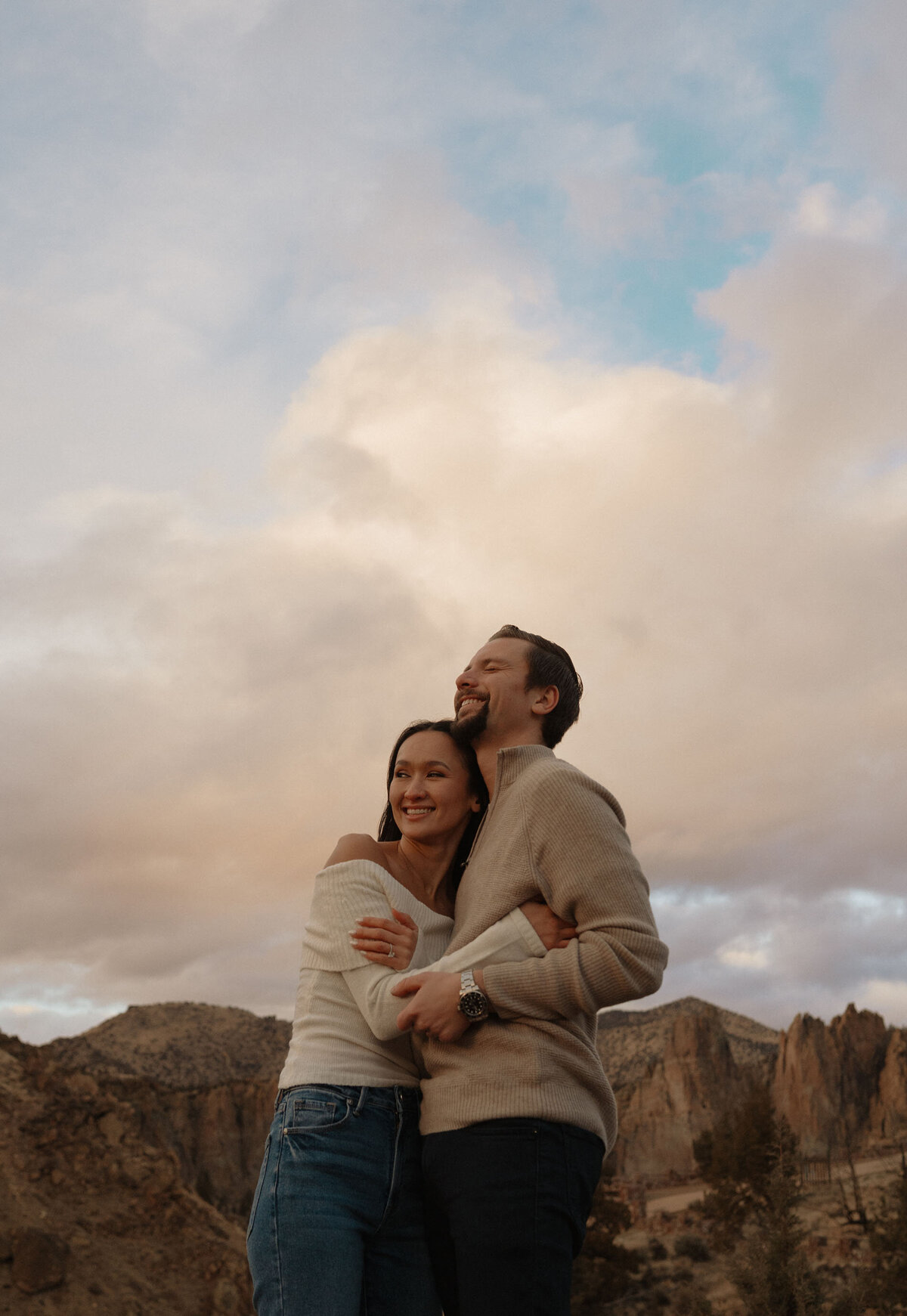 engaged couple embracing and smiling while standing against a cloudy sky and jagged rocks at Smith Rock State Park