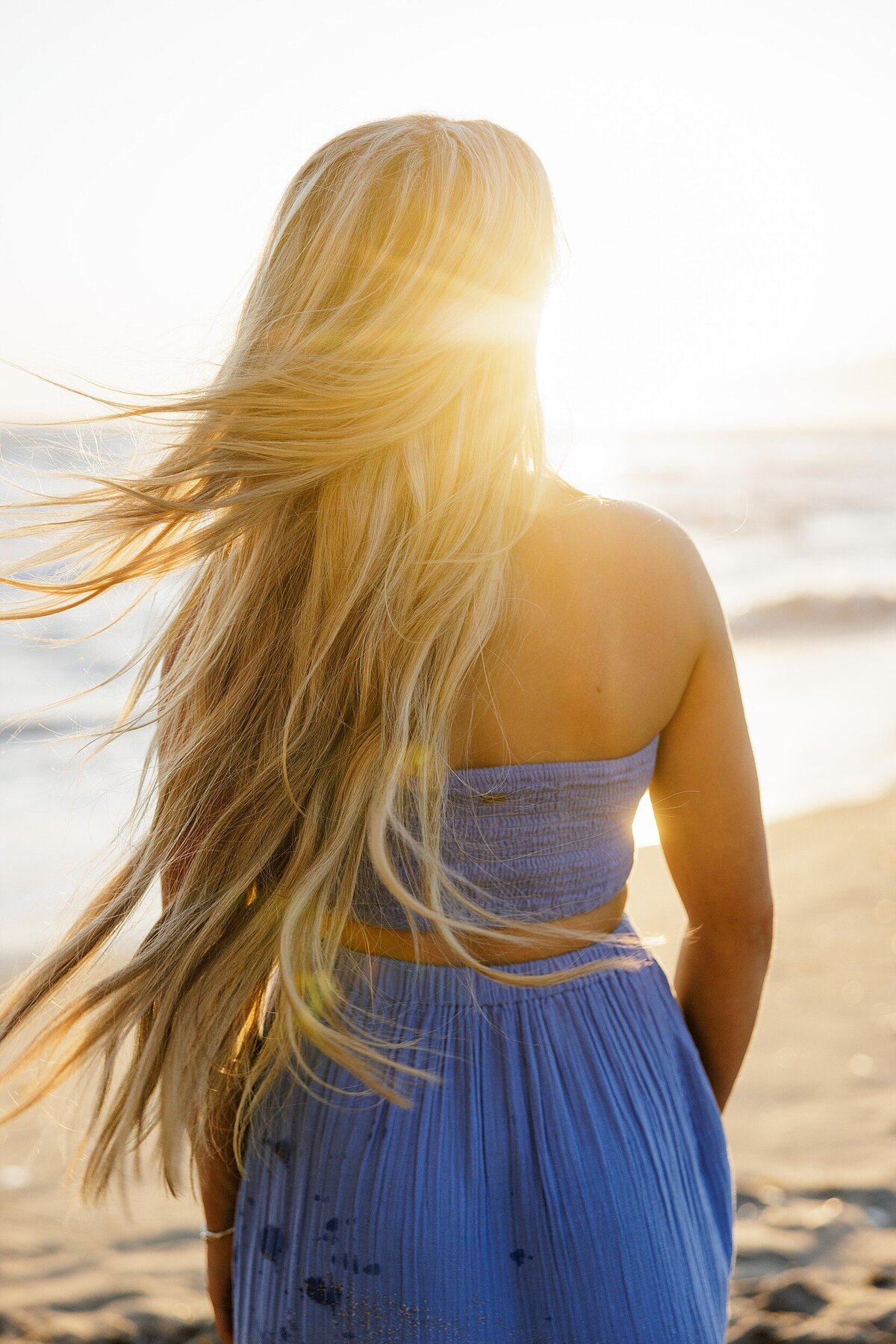 Golden hour senior photo of a blonde girl with long flowing hair walking along the beach in a soft blue two-piece dress, facing the sun