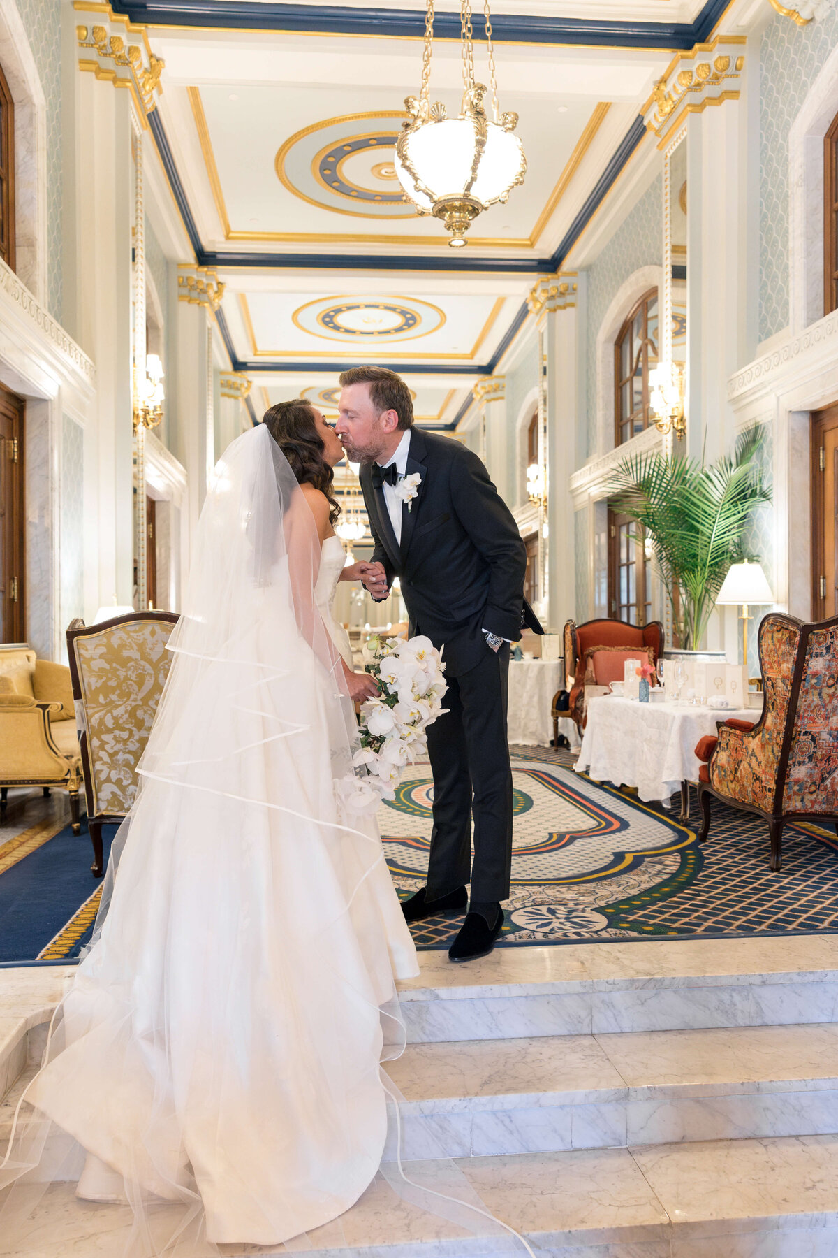A bride and groom smiling at each other. The groom wears a black suit with a bow tie, and the bride is in a white dress with a veil. They are in an elegant setting with flowers and soft lighting in the background.