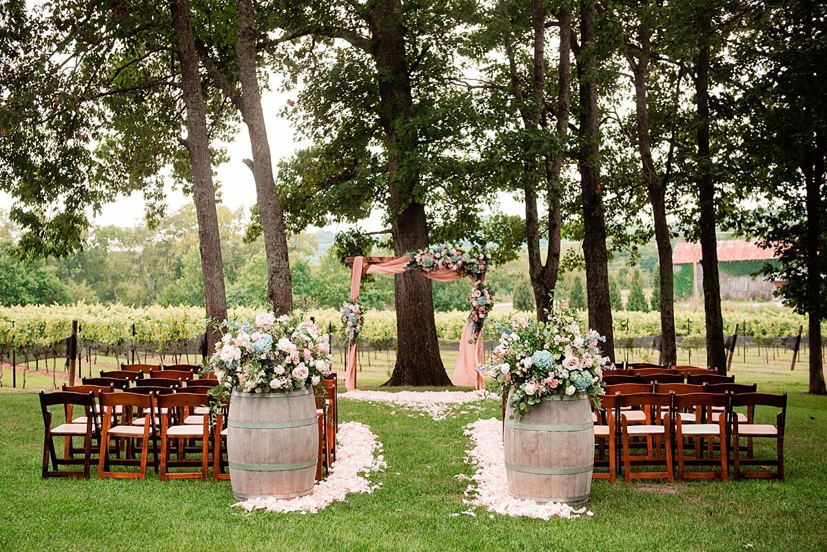 Arrington Vineyard Wedding  with brown fruitwood folding chairs with tan seats face several tall trees and rows of grapevines. On either side of the aisle are large wine barrels with large white and blush floral arrangements. The aisle is heavily lined with white and blush rose petals as it leads up to a wooden arbor with large sprays of roses and greenery.