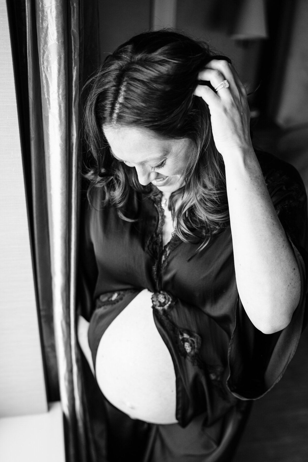 A pregnant woman stands by a window, gently cradling her bare belly while looking down with a serene expression. The black-and-white photo captures a quiet, reflective moment.