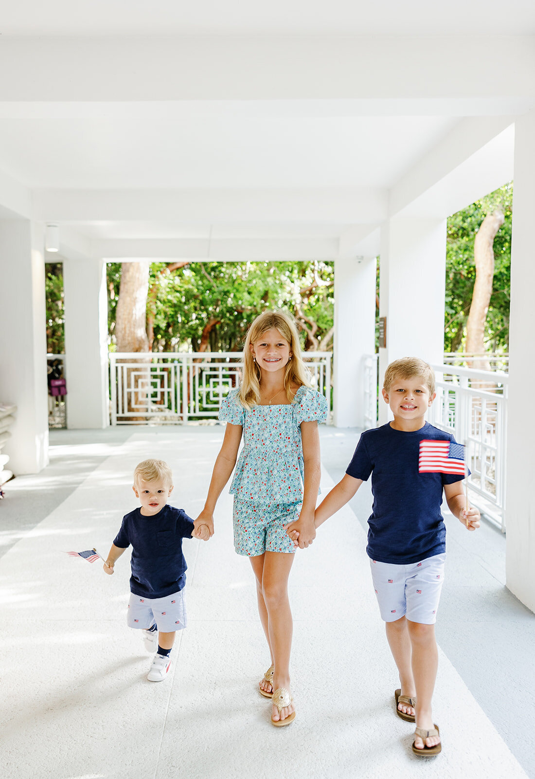 Three siblings hold hands as they walk together in a bright white corridor during a Florida Keys family session.