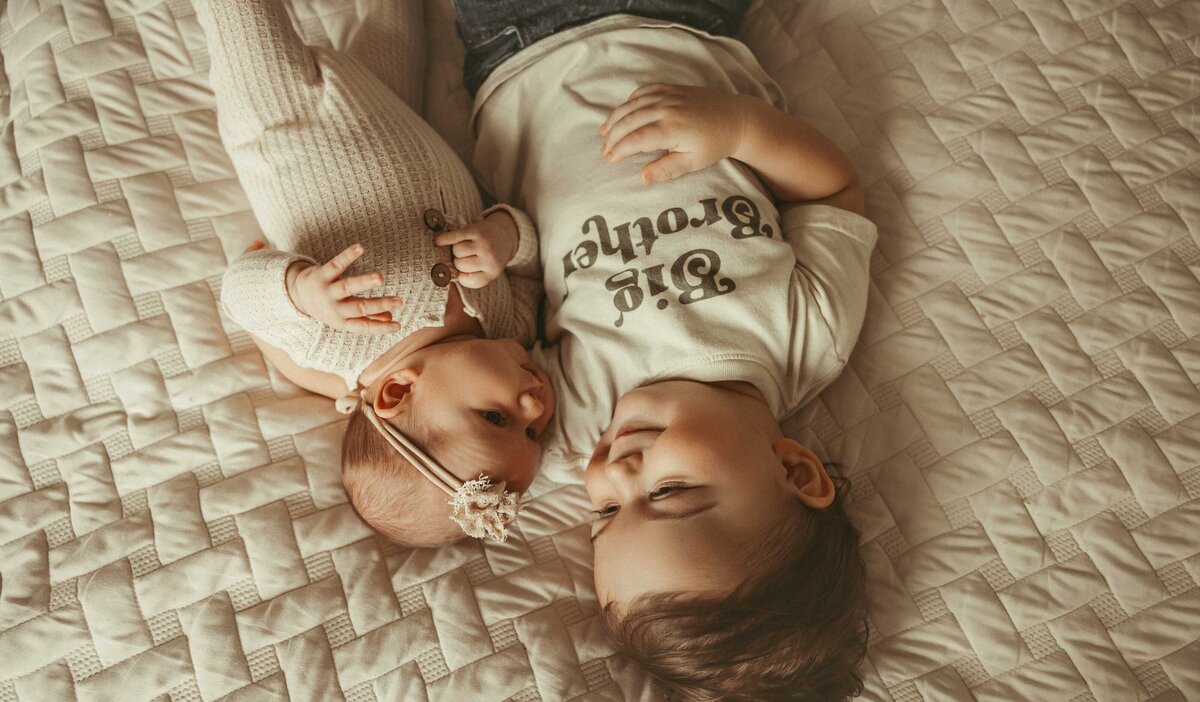 Young boy laying on a textured blanket wearing a big brother t-shirt. He is looking at his baby sister who is laying right by him