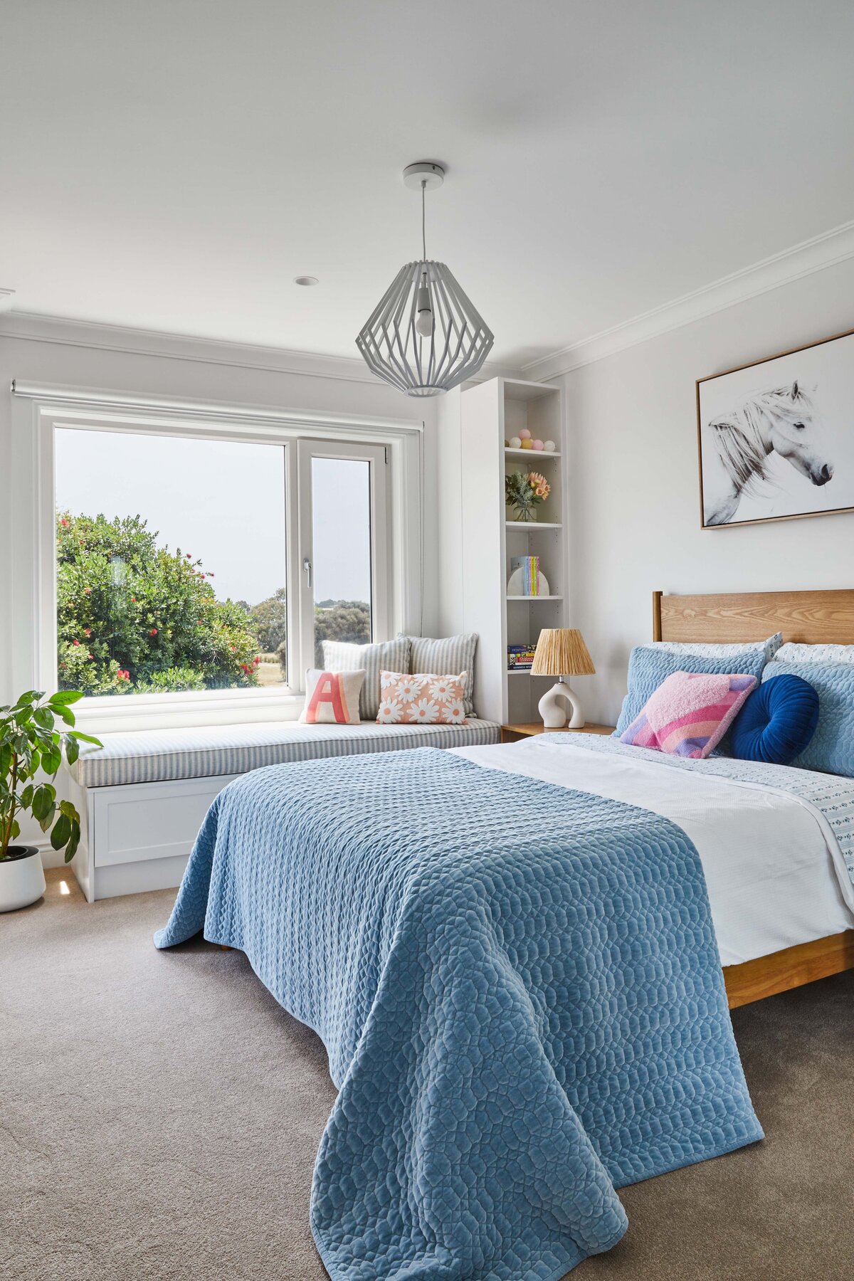 Bright bedroom with light timber bed, blue quilted bedding, colourful cushions and striped window seat with garden views.