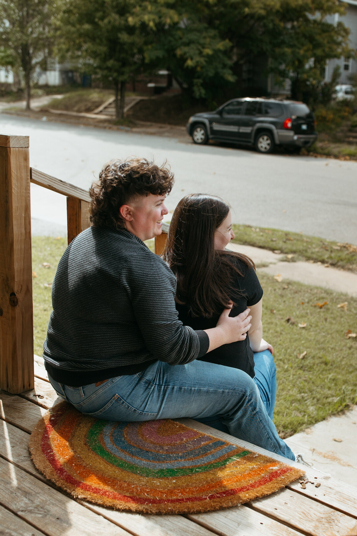 Two people sitting closely together on a colorful front porch mat, enjoying a quiet moment with a suburban street in the background