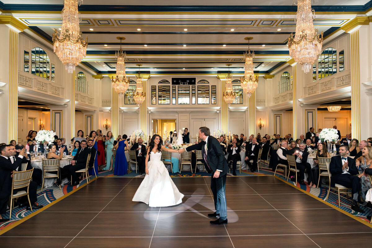 A couple dances in the center of a large, elegant ballroom with chandeliers and high ceilings. Guests surround the dance floor, sitting at round tables and watching the couple with smiles and applause.