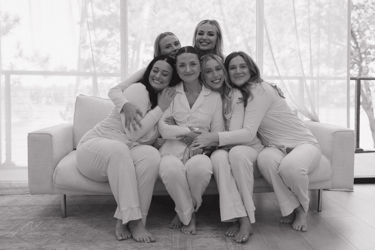 Bridesmaids in pink collared pajamas lean in giggling and smiling hugging the bride who is in white collared pajamas. There are 4 girls sitting on the main part of the couch and two girls up on the back rest of the couch crouching down. There is a wall of windows behind the couch covered in a slightly see through curtain.