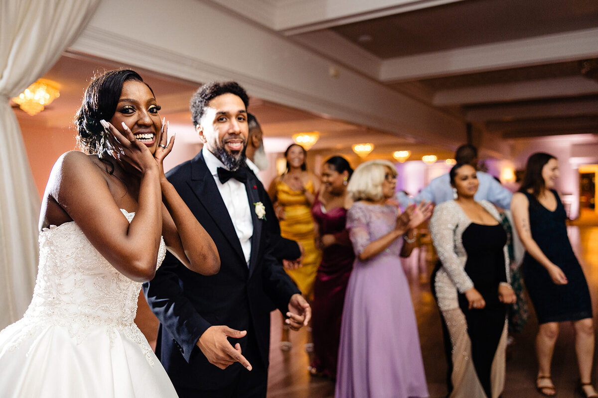 Bride in a white gown and groom in a tuxedo playfully dancing, with guests clapping in the background