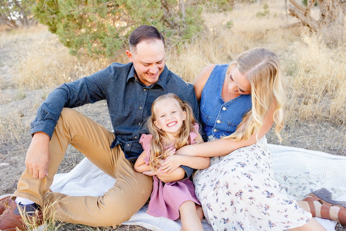 A little girl laughs as her parents tickle her while sitting on a blanket in the scenic Eagle Mountain, Utah, landscape. Their joyful expressions capture a carefree, happy moment.