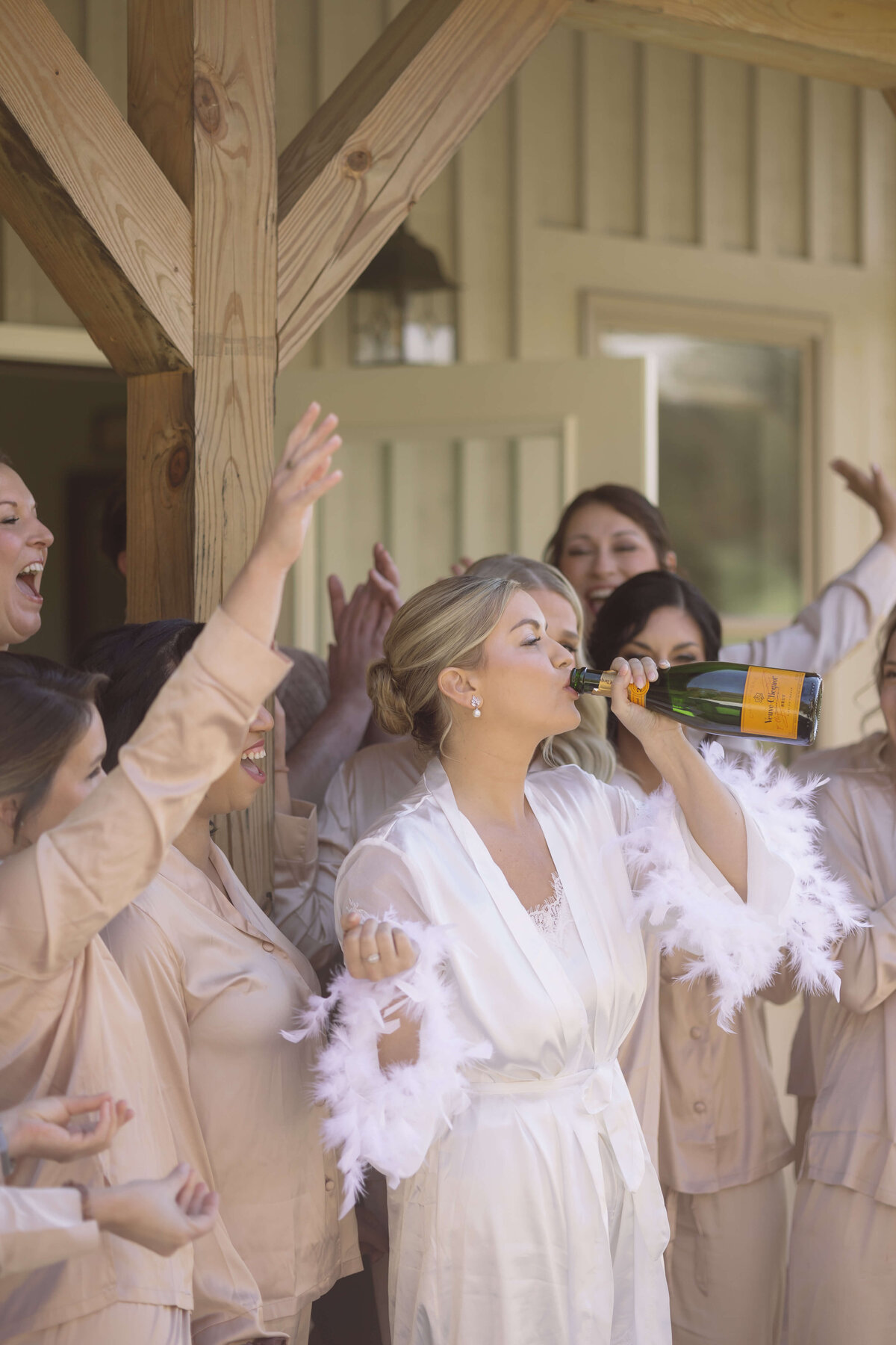 bride drinking champagne straight from the bottle while in her white robe with feather sleeves surrounded by her bridesmaids who are in blush pink pajamas photographed by Virginia wedding photographer