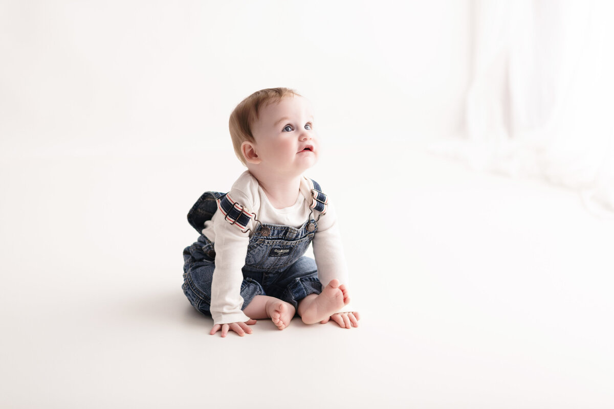 Clean milestone portrait of a baby in overalls with a white background—capturing childhood curiosity and innocence.