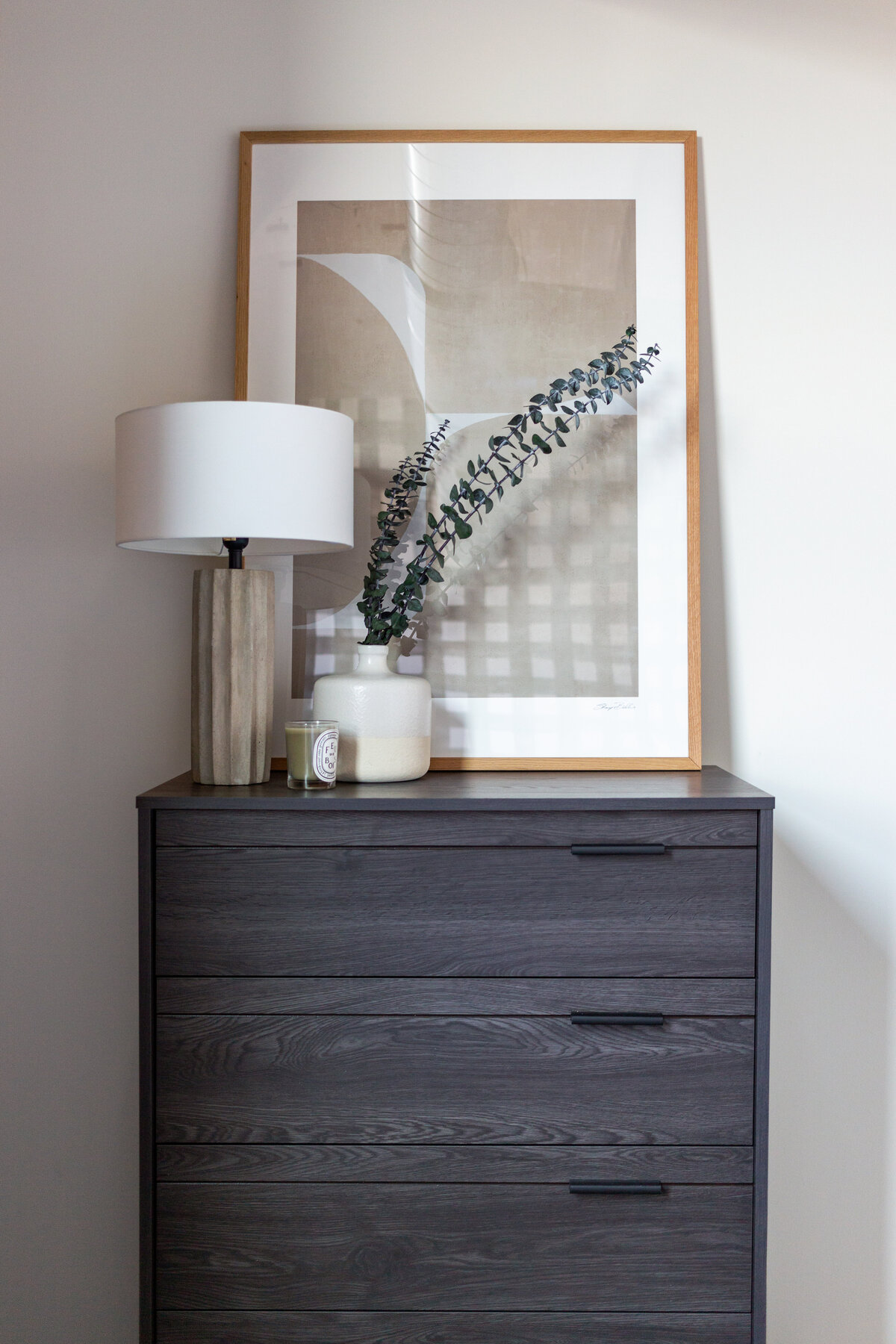 Black wooden dresser with scalloped concrete table lamp on top, white ceramic vase filled with eucalyptus and diptyque candle and abstract beige wooden framed artwork