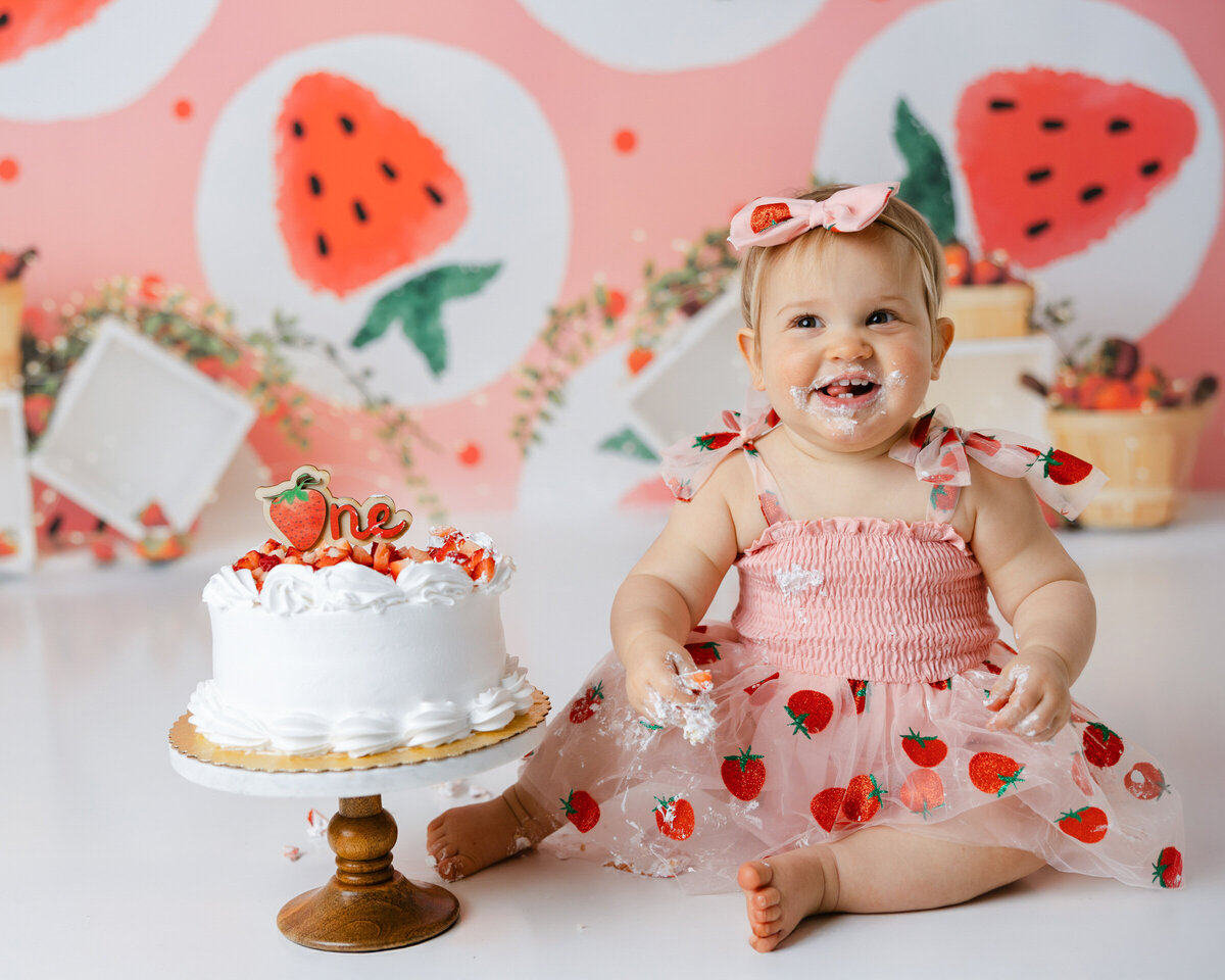 Baby in strawberry-themed dress enjoying cake smash with watermelon backdrop. Photograph by Yucaipa cake smash photographer Kaitlyn Dawn Photography.