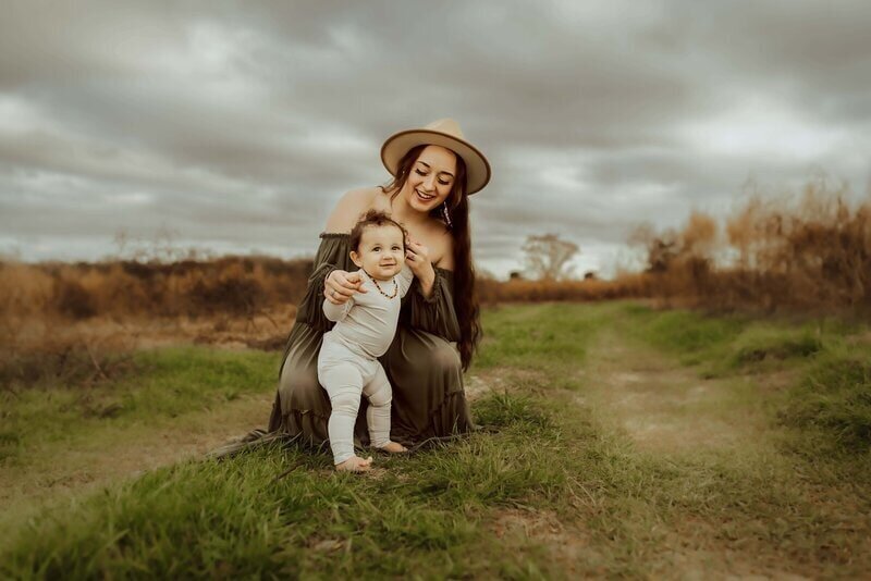 A woman in a hat crouches, holding a smiling baby on a grassy path under a cloudy sky.