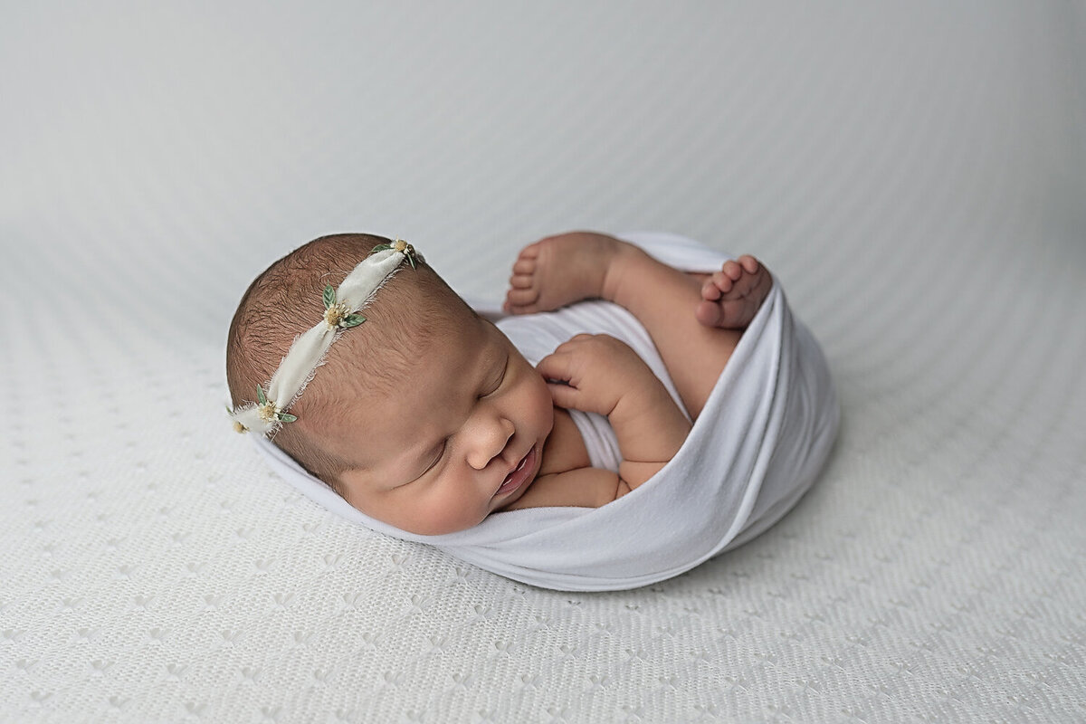 A newborn baby sleeps in an open white swaddle and a headband