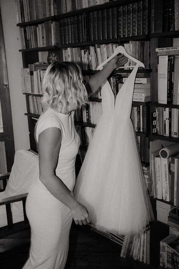 Gwladys, en robe blanche, observe sa robe suspendue devant une bibliothèque. Instant intime avant la cérémonie, capturé par Kamescop en Bretagne.