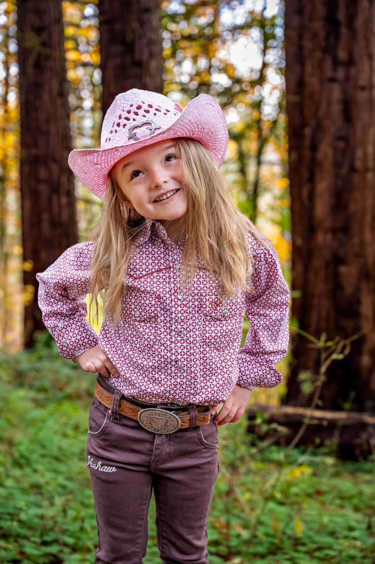 The image shows a young girl standing outdoors in front of blurred trees. She has long blonde hair and is smiling. She wears a pink cowboy hat with a heart-shaped decoration, a patterned long-sleeve shirt with red and white designs, brown pants with the word "Veehaw" embroidered on the left thigh, and a brown belt with a large oval buckle featuring a heart design. Her hands rest on her hips.