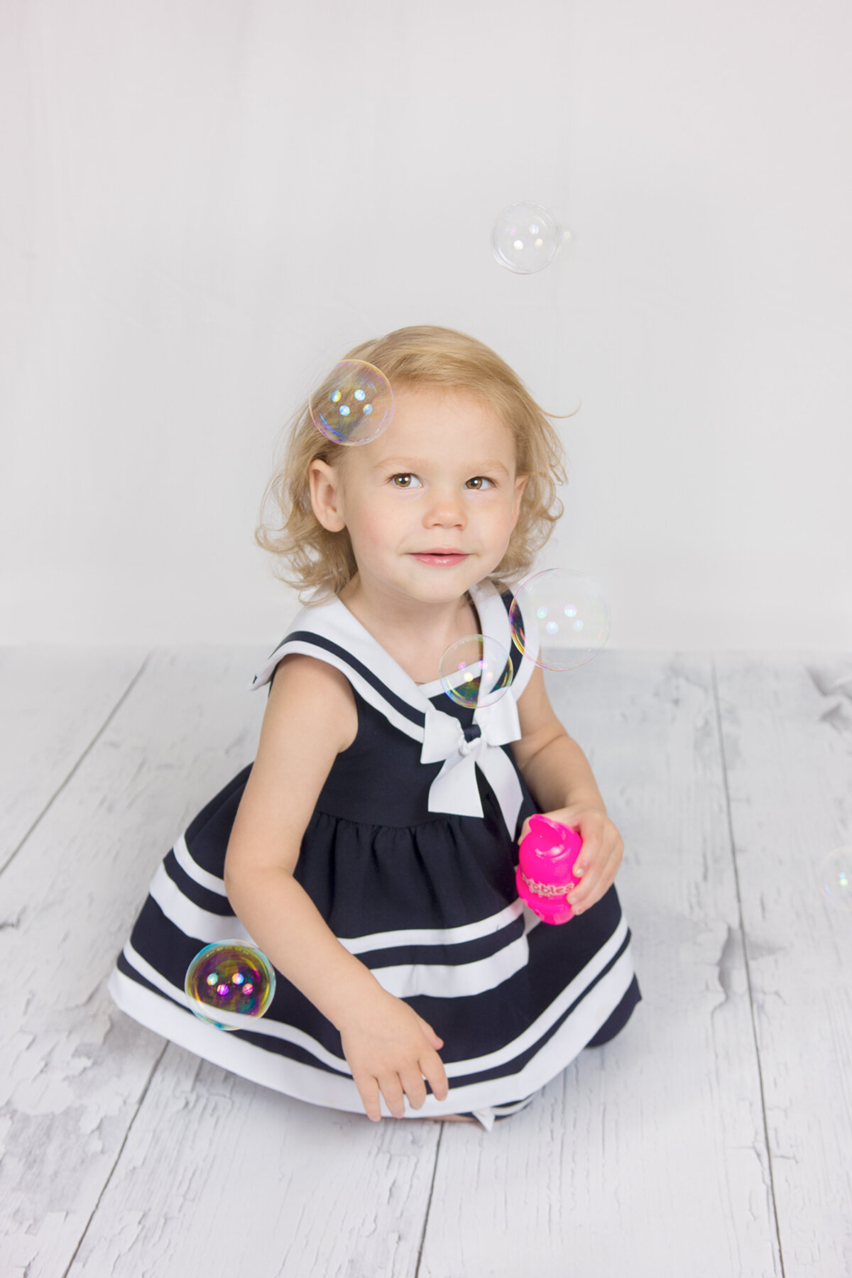 A toddler sits on a white hardwood floor and blows bubbles during a family studio-like photography session in McKinney, TX.