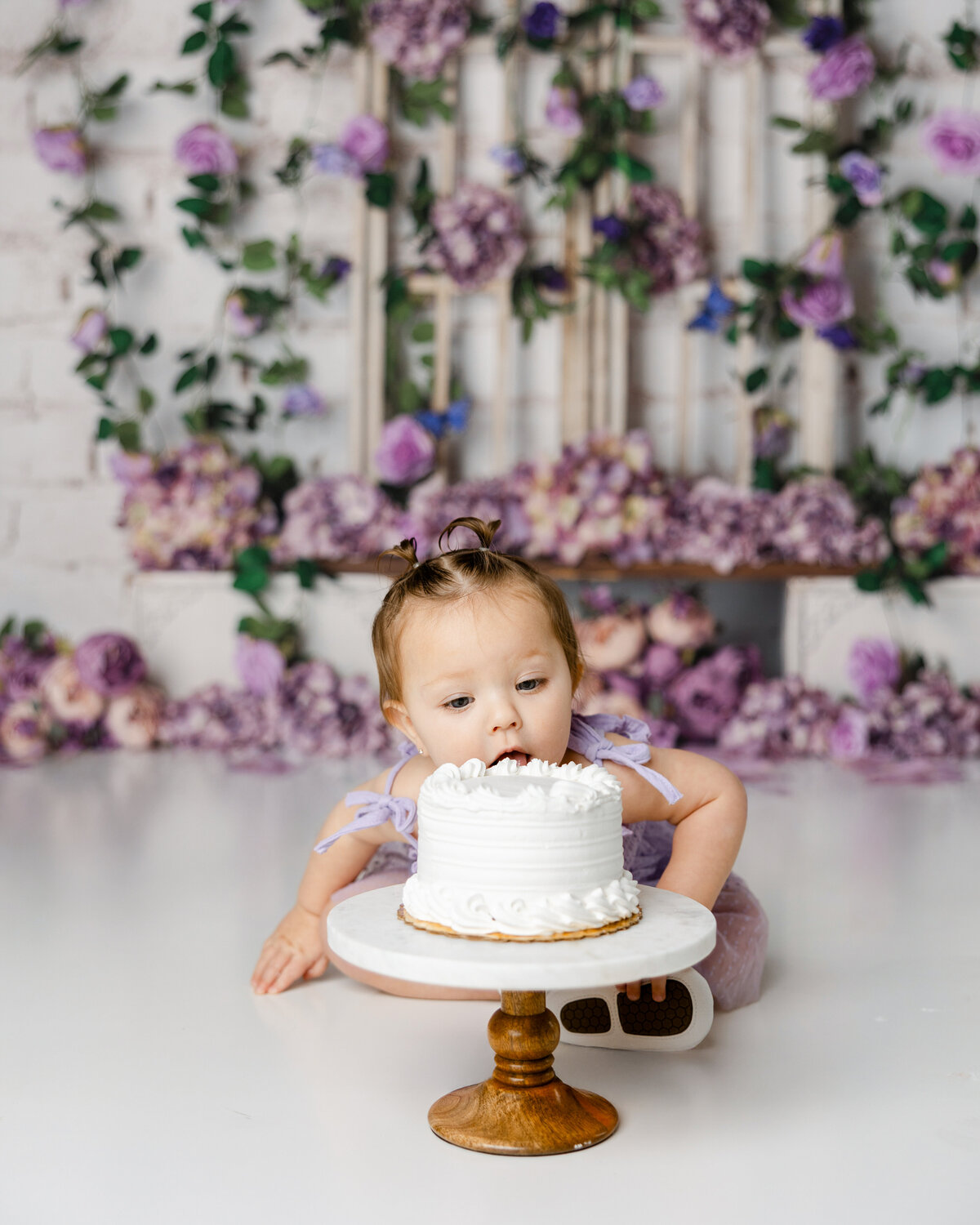 Baby leaning forward to taste a white cake during a cake smash session with a purple floral background. Photograph by Yucaipa cake smash photographer Kaitlyn Dawn Photography.
