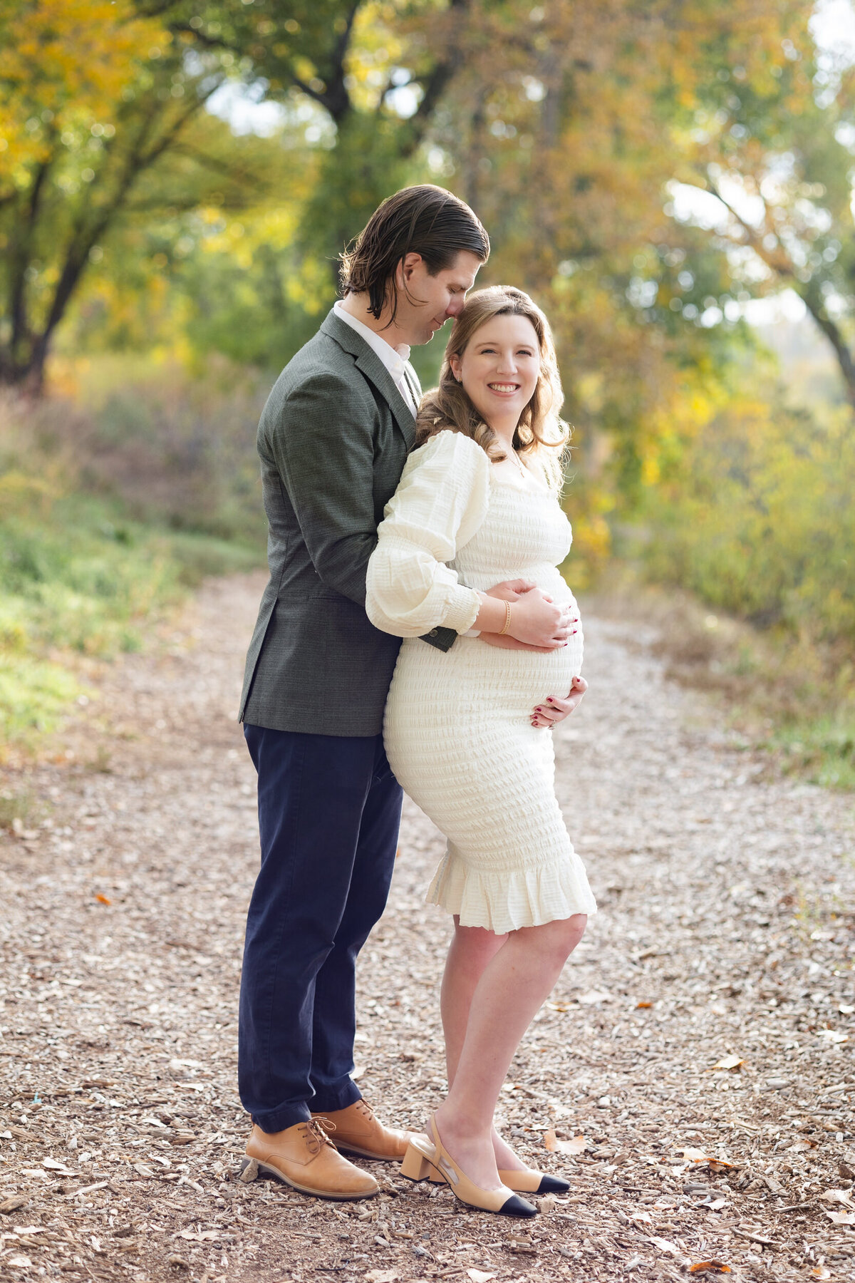 A man leans his head against his pregnant wife's head as he hugs her from behind and she hold her belly with both hands and smiles at the camera.