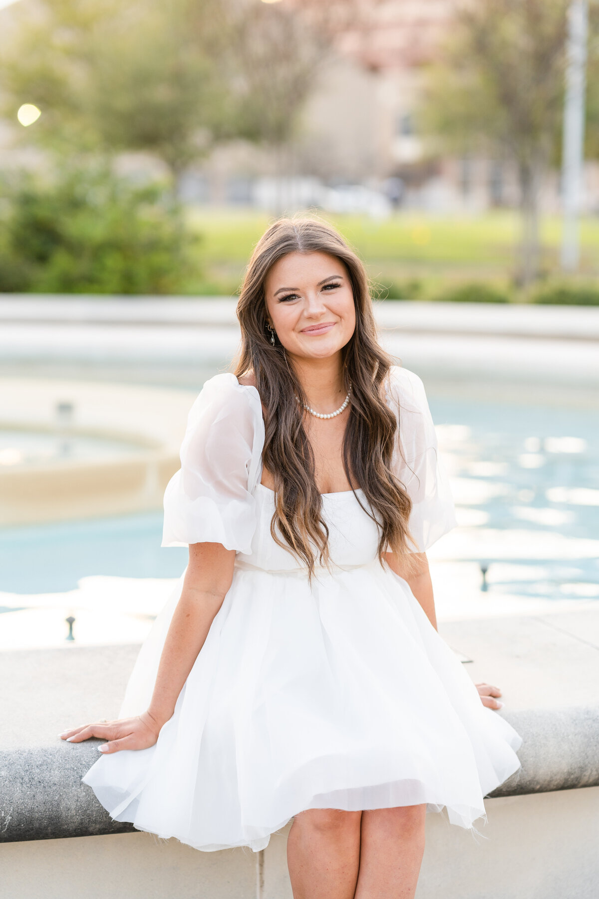 Texas A&M senior girl sitting on side of fountain wearing a puffy white dress and smiling in front of Kyle Field