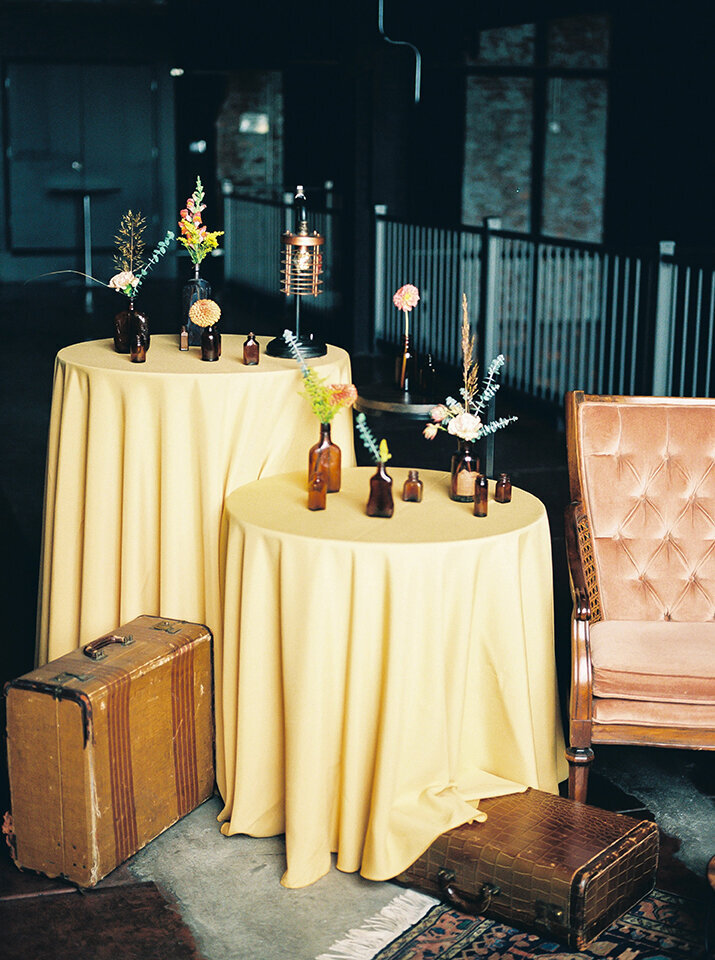 Two rounded tables with yellow table cloths and amber bottles filled with flowers next to blush-colored chair and suitcases.