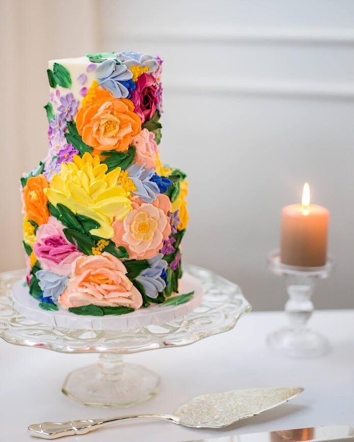 A two tier cake sits on a clear cake stand with a candle in the background. The cake is covered with buttercream painted florals in bright rainbow colors. 
