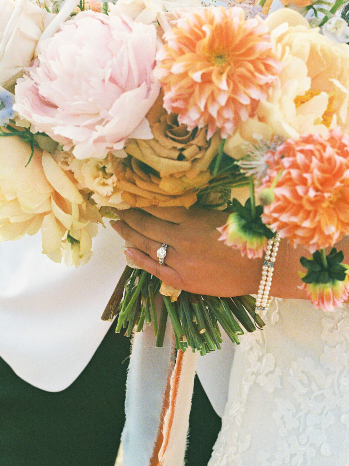 Bride holding a bouquet of pastel flowers, featuring pink and orange blooms. Her hand showcases a diamond ring and pearl bracelet, conveying elegance and joy.