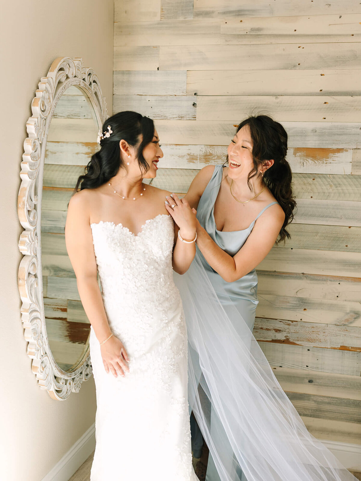 Bride and bridesmaid share a joyful moment; one in a lace wedding gown and the other in a light blue dress, standing near a rustic wooden wall and vintage mirror.