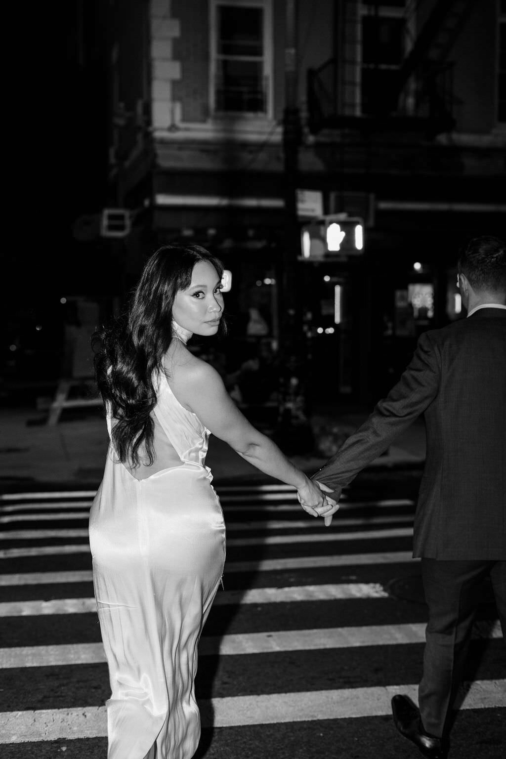 black and white photo of a woman looking over her shoulder as she holds hands with her groom and walking across a city street