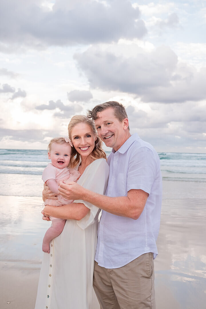 Light-filled family of 3 at a Brisbane beach: Lifestyle portrait with mum and dad holding baby