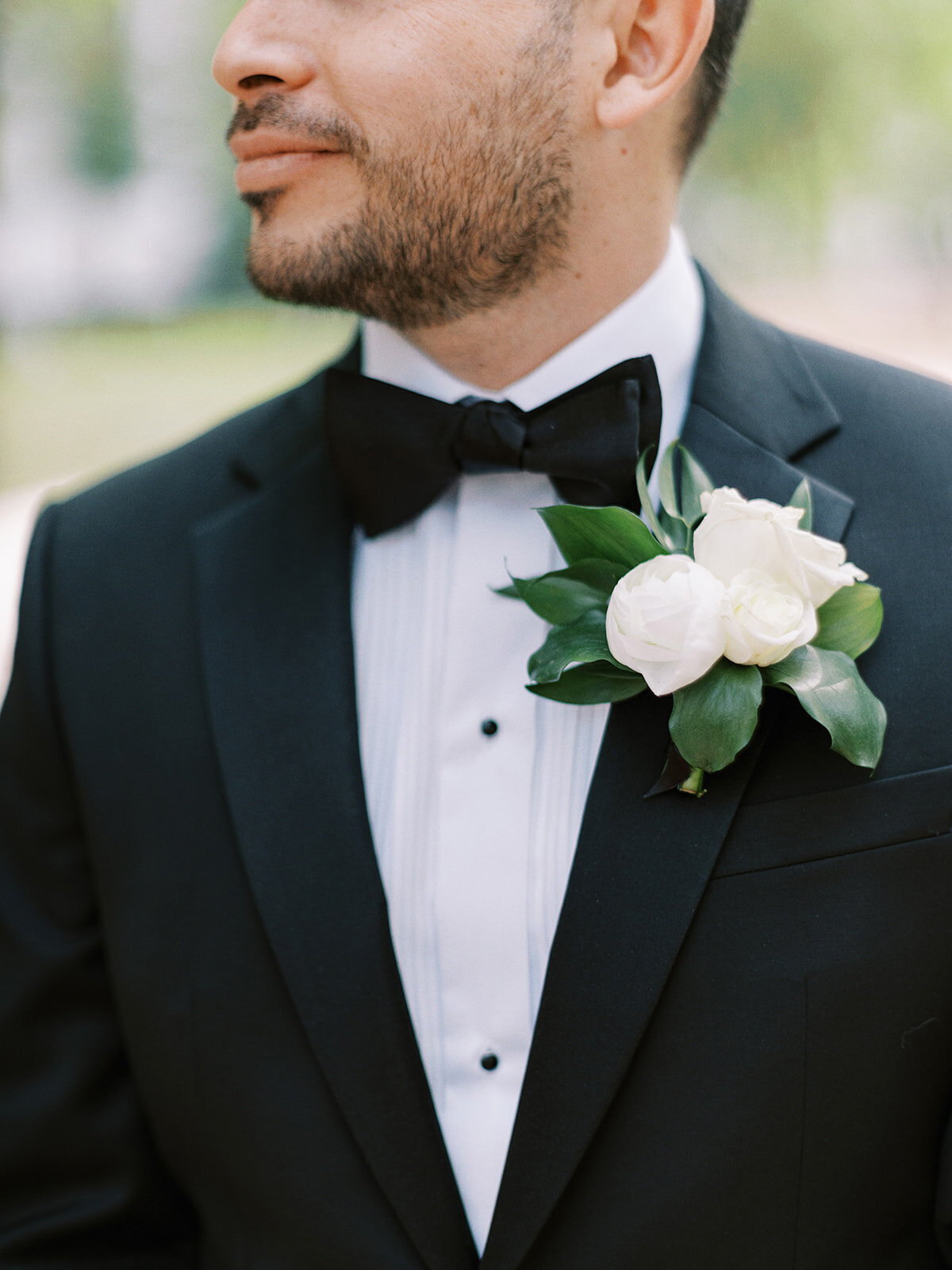 Man in a black tuxedo with a white dress shirt and black bow tie at a cultural Calgary wedding. He has a beard and is wearing a boutonniere with white flowers and green leaves on his lapel. His head is turned to the side, capturing the elegance of the Fairmont Palliser Wedding in Calgary.