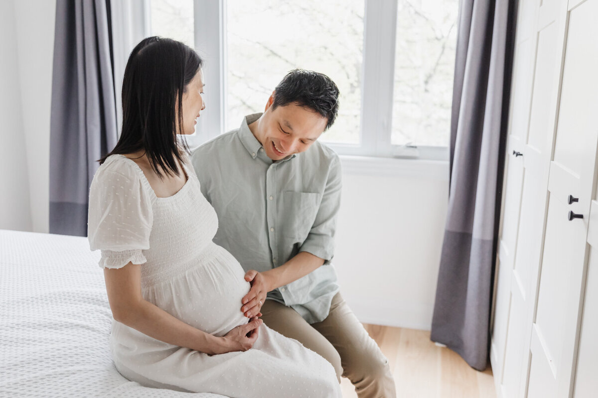 Husband gently touching pregnant wife's belly. They are sitting on the edge of a white bed with a big window behind them.