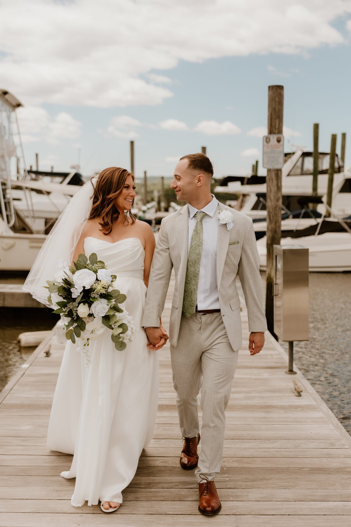 Bride and groom walk hand in hand on a marina dock during their elegant Westchester wedding, smiling at each other with boats in the background.