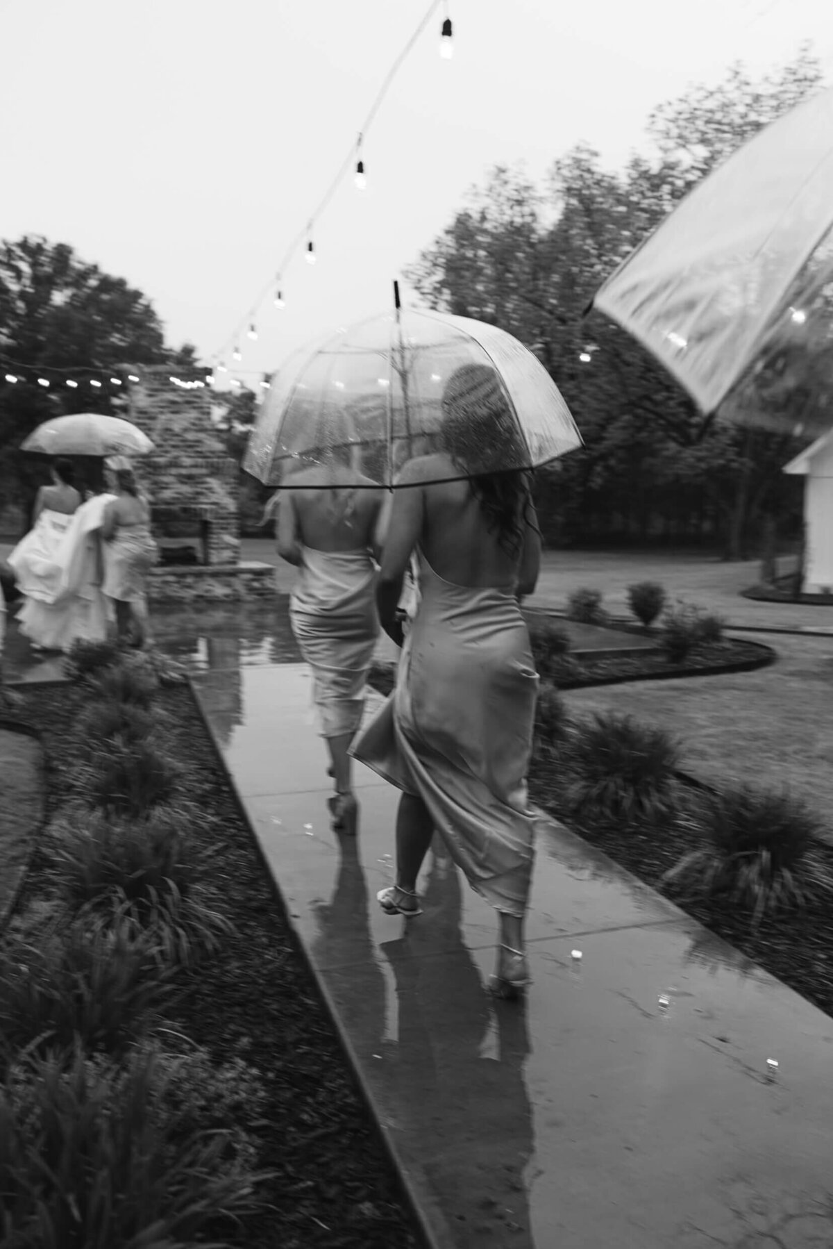 A black and white wedding photo of the bridesmiads walking to the ceremony holding umbrellas as it is raining