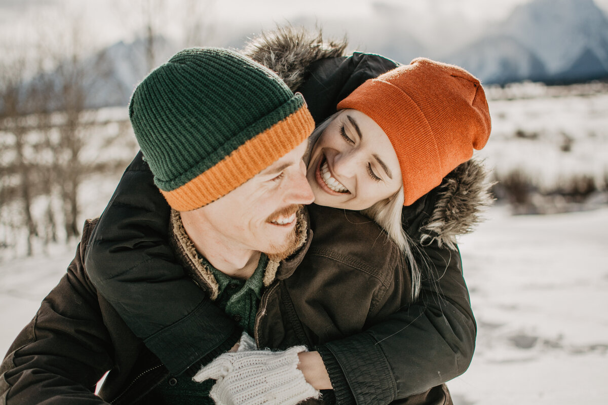 Winter engagement session with woman leaning over her fiances shoulder and smiling at him as he smiles as her , both are wearing beanies and winter gear