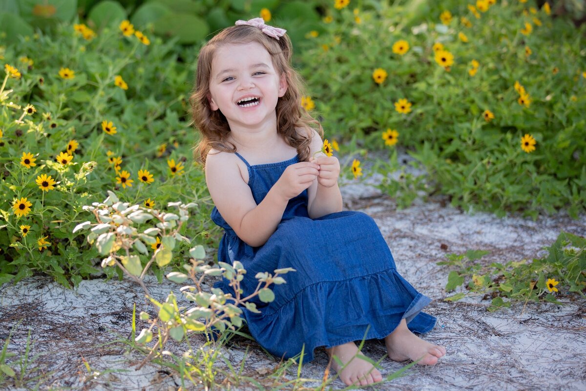 Little girl in a blue dress laughs for the Anna Maria Island family photographer with yellow flower background