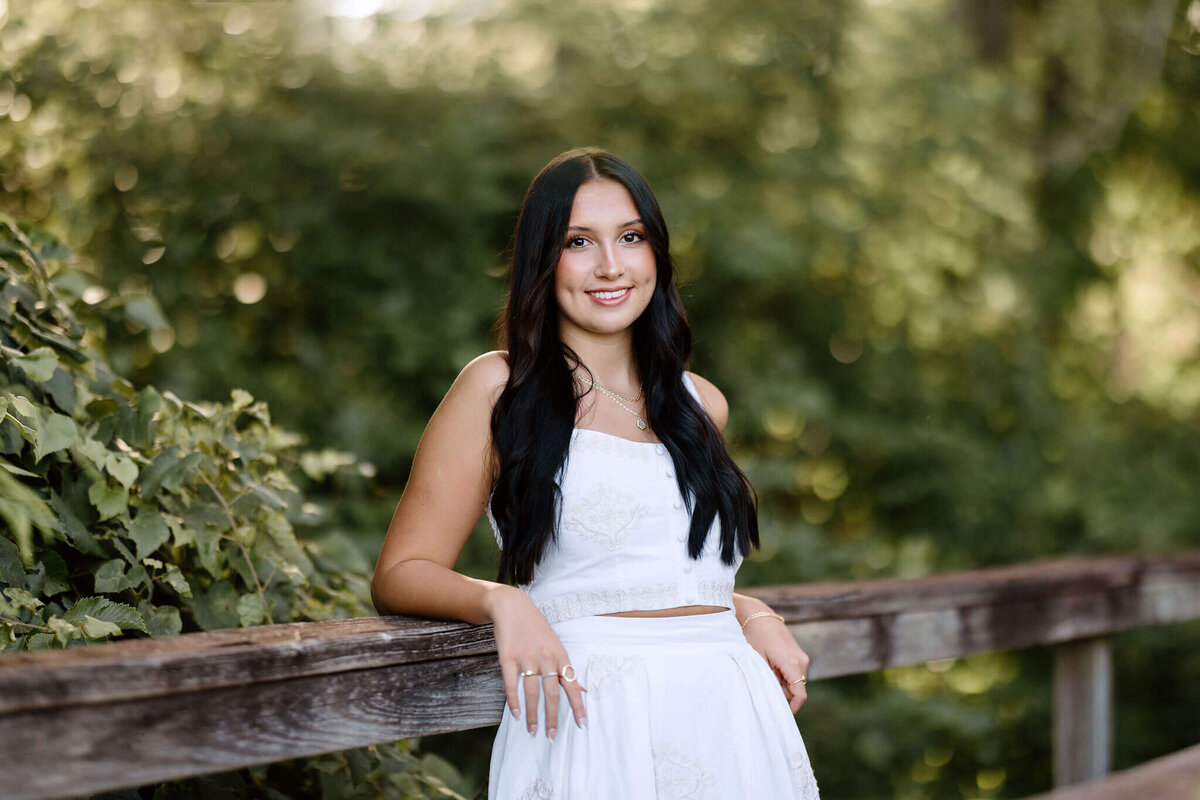 High school graduate wearing white long dress leaning against bridge 