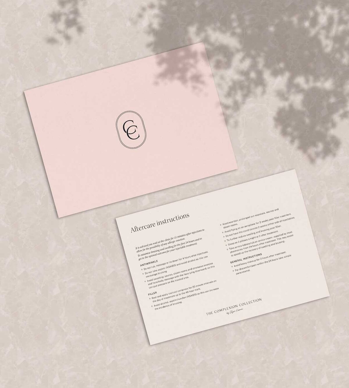 Flatlay of The Complexion Collection Aftercare Instructions card showing front and back design, pastel pink and monogram submark design on front and white with information on back sitting on a marble background with flower shadows overlayed